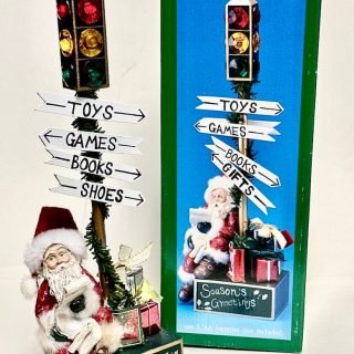 Large decorative flashing stop-light Santa sitting on a base, checking his list with presents at the base of a traffic signal and signs pointing different directions for gifts.